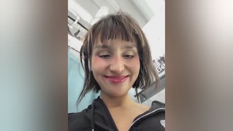 Media: Video of a young woman with light brown skin, short black hair with bangs, wearing a black jacket with a white logo, smiling, in a brightly lit, white-walled room with medical equipment in the background.