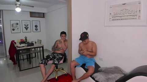 Media: Video of two shirtless men in casual shorts, one with tattoos, sitting on a couch in a sparsely decorated, modern living room with minimalist art, city map, and a ceiling fan.