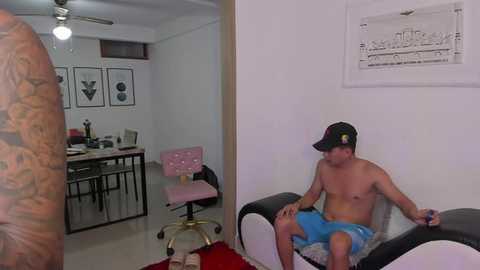 Media: Video of a shirtless, tattooed man with a cap, lounging on a black and white bean bag chair in a minimalistic, white room with a desk, chair, and cityscape art on the wall.