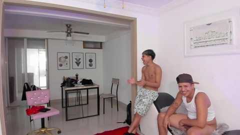 Media: Video of a modern living room with two shirtless men in swim trunks, one wearing a cap, playing video games.