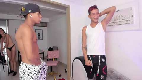 Media: Video of two men in casual clothes: one shirtless in patterned shorts, the other in a white tank top with floral shorts. Background shows a modern, minimalist apartment with white walls and furniture.