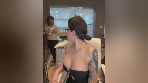 Media: Video of a woman with dark hair in a bun, wearing a black strapless dress, showing off a tattooed arm, seated at a table. A man in the background talks to another person.