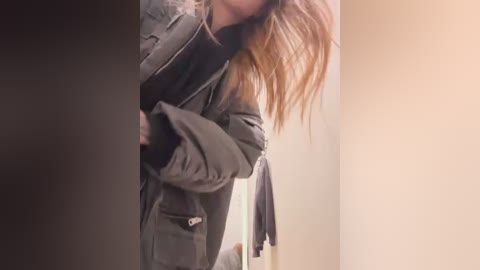 Media: Video of a person with long, wavy brown hair, wearing a dark jacket, standing in a beige-walled room with a towel hanging on the left. The image is slightly blurry and dimly lit.