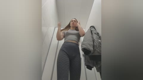 Media: Video of a tall, slender, Caucasian woman with long, straight hair, wearing a grey crop top and high-waisted, patterned leggings. She stands in a narrow, white-tiled hallway, reaching up, possibly adjusting her hair. A grey jacket hangs on a hook to the right.