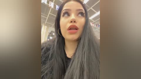 Video of a woman with long, straight, silver hair, wearing a black dress, and hoop earrings, standing indoors with a neutral background.