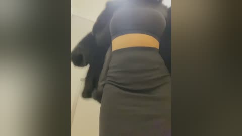 Media: A video of a woman in a tight, dark gray dress with a wide, mustard-colored waistband, taken from a low angle, showing her full figure and back. The background is blurred, featuring light-colored walls.