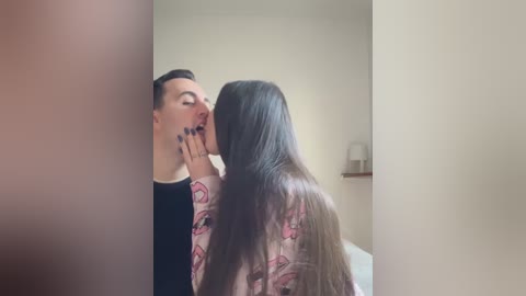 Media: Video of a man and woman in an intimate moment. The man, with short hair and tattoos on his face, is kissing the woman, who has long hair and is wearing a pink shirt. The background is a minimalist bedroom with beige walls and a small shelf.