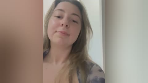 Media: Video of a young Caucasian woman with long blonde hair, wearing a floral-patterned blouse, standing indoors with a neutral expression.