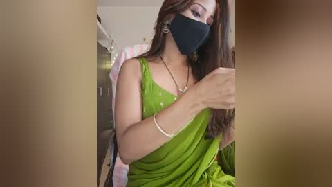 Media: Video of a South Asian woman with medium skin tone and long brown hair, wearing a green saree, black face mask, and gold jewelry, sitting indoors, possibly in a bathroom.