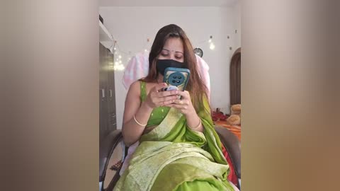 Media: Video of a young woman in a green sari, taking a selfie indoors with a blue phone, wearing a black mask. Background features a bed with stuffed toys, white walls, and a wooden armoire.