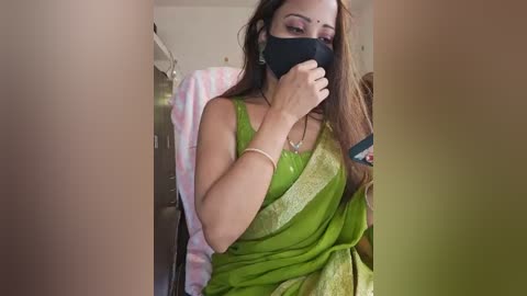 Media: A video of a South Asian woman with long brown hair, wearing a green saree, black mask, and a gold bracelet, sitting in a hospital room, holding a phone.