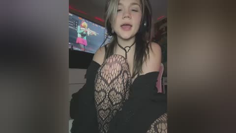 Media: Video of a young woman with long, black and blonde hair, wearing a black fishnet top and a black off-shoulder jacket, standing in a dimly lit room with a TV screen showing a cartoon character.