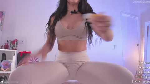 A video of a curvy woman with long black hair, wearing a beige sports bra and leggings, sitting on a bed, holding a water bottle.