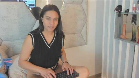 Media: Video of a woman with long black hair, wearing a black sleeveless top with white trim, sitting on a beige couch, typing on a laptop. Background includes a beige geometric-patterned wall and a white shelving unit with hair products.