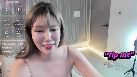 Media: Video of an Asian woman with light skin, shoulder-length brown hair, and a slender figure. She wears a light-colored, sleeveless top. Background shows a modern room with a white door, white walls, and a \"tip me\" sign.