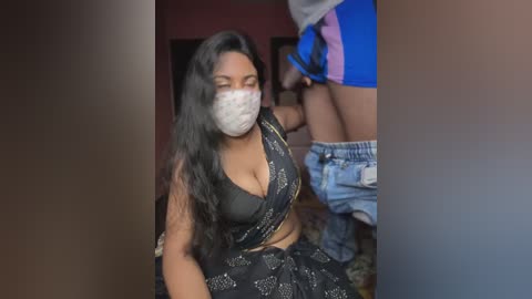 Media: Video of a South Asian woman with long black hair, wearing a black crop top and floral skirt, gas mask, and jeans, standing in a dimly lit room.