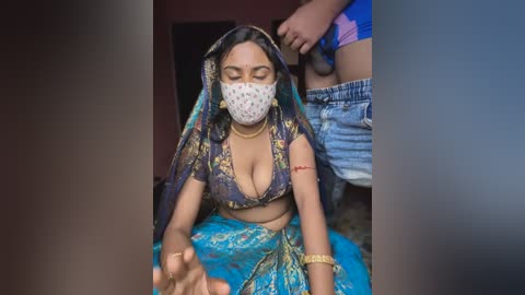 Media: Video of a South Asian woman in traditional attire, wearing a colorful sari and face mask, sitting on a floor. A partially visible person in blue shorts stands behind her.