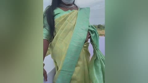 Video of a woman in a yellow-green sari, accessorized with a necklace, holding a green cloth. Background features a white wall and a blurred, natural landscape.