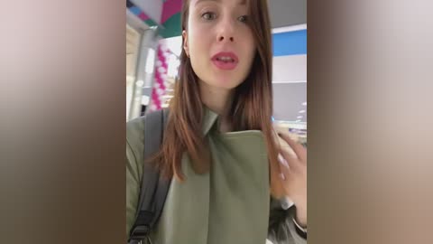 Media: Video of a young Caucasian woman with straight, shoulder-length auburn hair, wearing a olive-green shirt and gray backpack, indoors with a blurred background featuring colorful banners and a window.