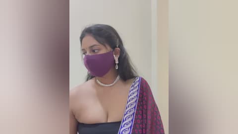 Media: Video of a South Asian woman with medium-dark skin, wearing a black strapless top, a purple mask, and a red sari with white pattern, in an indoor setting.