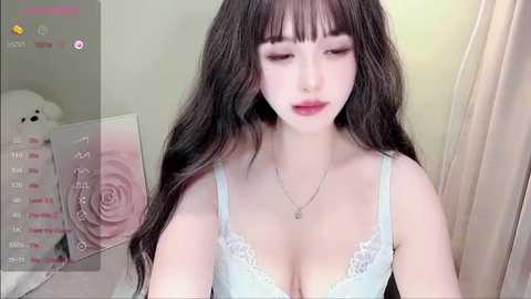 Media: A video of a fair-skinned woman with long, wavy dark brown hair, wearing a light blue lace bra and a delicate necklace, sitting in a soft-lit room with a white bear and a rose-themed calendar in the background.