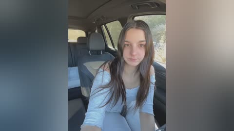 Media: Video of a young Caucasian woman with long brown hair, wearing a white off-shoulder top, seated in the back seat of a car with black leather seats. The background features a blurred, natural outdoor scene.