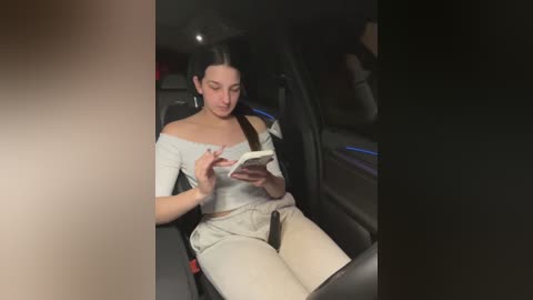 Media: Video of a woman with long dark hair, wearing a white off-shoulder top and beige pants, reading a book in a car.