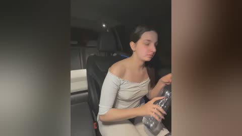 Media: Video of a Caucasian woman with dark hair, wearing a white off-shoulder top, seated in a modern car. She is holding a water bottle, with a blurred man's arm and torso in the foreground.