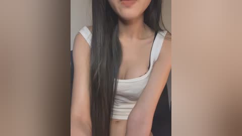 Media: A video of an Asian woman with long, straight black hair and a pale complexion, wearing a white tank top that reveals her cleavage. She stands against a beige wall, partially cropped at the top and bottom.