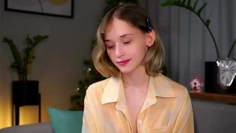 Media: Video of a young woman with light skin and shoulder-length blonde hair, wearing a yellow tie-dye shirt, smiling softly. Background features indoor plants, a potted cactus, and a cozy, dimly lit room with a fireplace.