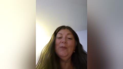 A blurry video of a middle-aged woman with long dark hair, wearing a black jacket, smiling and making a funny face against a white wall.