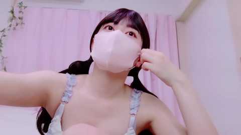 Media: Video of an East Asian woman with long black hair, wearing a light blue floral-patterned bra, pink face mask, and a casual expression, indoors with light pink curtains and white walls in the background.