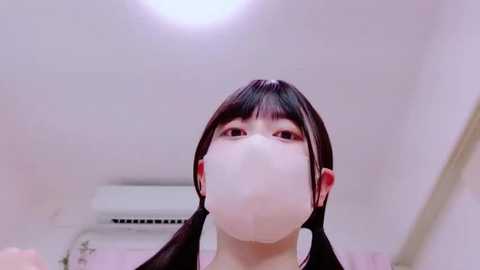 Media: Video of an Asian woman with long black hair and bangs, wearing a white face mask, standing indoors with an air conditioner visible behind her.