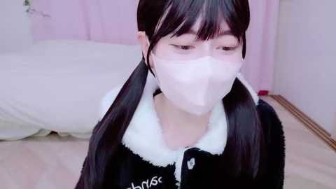 Media: A video of a young Asian woman with long black hair in pigtails, wearing a white face mask, black fur-trimmed coat, and standing indoors.