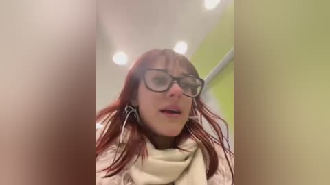 A video of a young woman with long, straight red hair, wearing black-framed glasses and a beige scarf, standing in a brightly lit, modern restroom with a green wall and white ceiling.