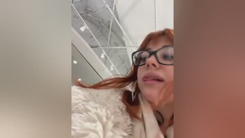 Video of a fair-skinned woman with long red hair, wearing black-framed glasses, looking down at a white fluffy rug in a modern, glass-enclosed room with a ceiling of exposed metal beams.