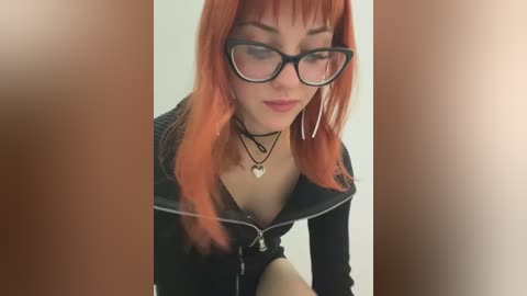 Video of a young woman with long, bright orange hair, wearing black-framed glasses, a black zip-up jacket, and multiple necklaces. She has fair skin and is indoors against a plain background.