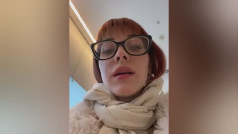 Media: Video of a Caucasian woman with short red hair, wearing black-rimmed glasses, a beige scarf, and a light sweater. The background is a modern interior with soft lighting and beige walls.