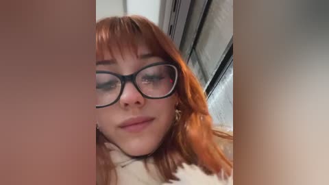 Media: Video of a young woman with fair skin and reddish-brown hair, wearing black-rimmed glasses and a white sweater. She has a neutral expression, with a blurred background of a tiled wall.