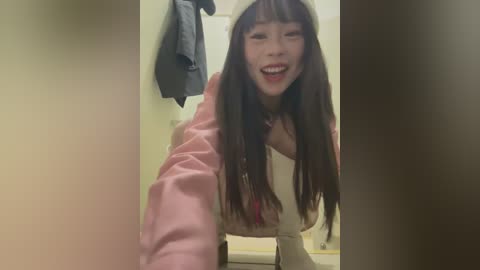 Media: Video of a smiling East Asian woman with long brown hair and bangs, wearing a pink jacket and white top, in a beige room with a gray coat hanging on the wall.