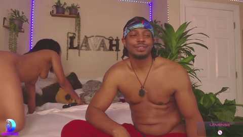 Video of a shirtless, muscular Black man with a headband, sitting on a bed with a guitar player in the background. The room is dimly lit with purple LED strip lights.