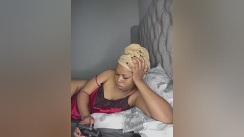 Media: Video of a Black woman with braided hair, wearing a red and black camisole, lying on a bed, holding a phone, head in hand, in a dimly lit room with gray walls and a patterned headboard.