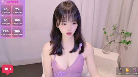 Media: A video of an Asian woman with straight black hair, wearing a purple lingerie top, sitting on a white chair in a minimalist room with a plant and curtains.