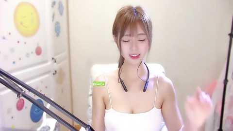 Media: Video of a young Asian woman with fair skin and long brown hair, smiling in a white tank top, wearing headphones in a cozy, brightly decorated bathroom with a white bathtub and colorful wall art.