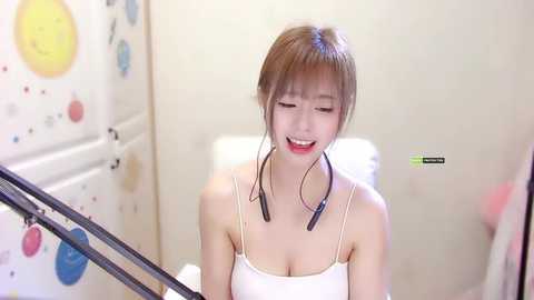 Media: Video of a young East Asian woman with light skin, brown hair, and bangs, wearing a white spaghetti-strap top, smiling. She has a headset on. Background features a playful, colorful wall with circular patterns.