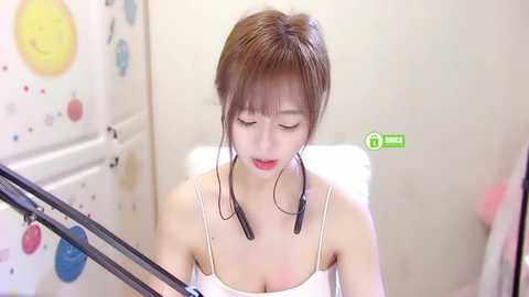 Media: Video of an East Asian woman with straight brown hair and bangs, wearing a white spaghetti-strap tank top, singing into a microphone, with a soft, pastel-colored background.