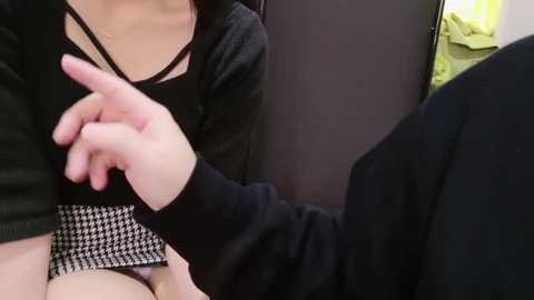 Media: Video showing a close-up of a woman's torso in a black top, with a hand gesturing and a checkered skirt visible. Background includes a yellow garment and a dark surface.