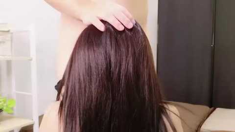 A video of a woman's long, straight, dark brown hair being massaged by a hand in a salon setting. The background includes a beige wall and a white shelf with green plants.