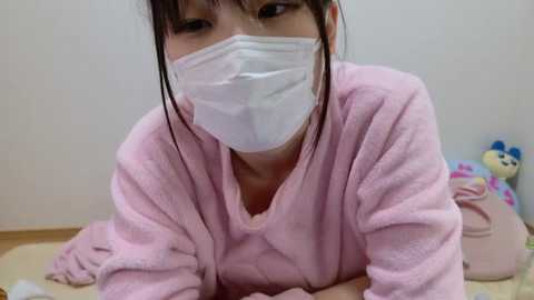 Media: Video of an Asian woman in a pink bathrobe, wearing a white surgical mask, lying face down on a bed with a plush toy beside her.