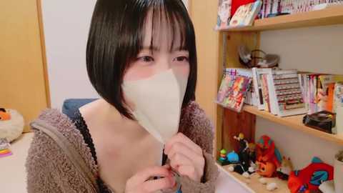 Media: Video of a young Asian woman with a bob haircut, wearing a beige knit cardigan, holding a large, white fan in front of her face. Background features a bookshelf filled with colorful toys and books.